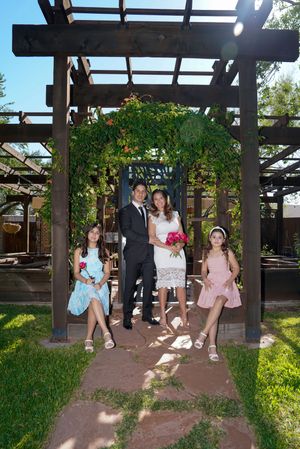 Couple in wedding attire with two girls in dresses posing under a wooden pergola with greenery outdoors.