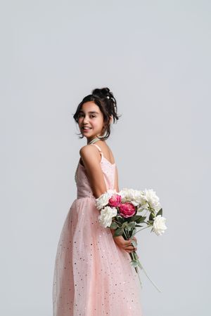 Young girl in pink tulle dress holding white and pink peonies bouquet, smiling over shoulder on gray background.