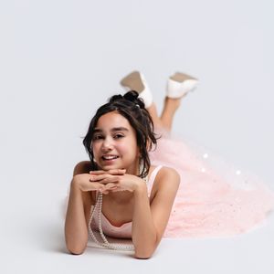 Young girl in pink tulle dress and pearl necklace poses lying down, smiling, wearing white heels on gray background.