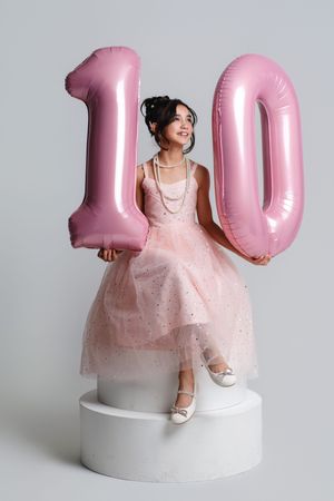 A smiling girl in a pink tulle dress holds large pink number 10 balloons while sitting on a white pedestal.