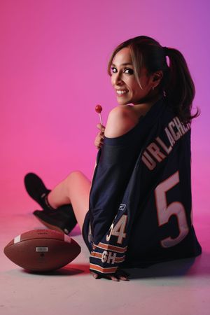 Woman in Chicago Bears jersey holding lollipop poses with football on pink background.