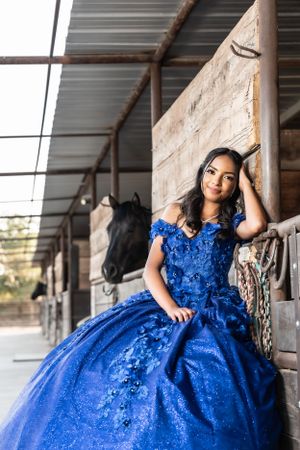 A young woman in a sparkling royal blue quinceañera gown poses elegantly in a rustic stable setting.