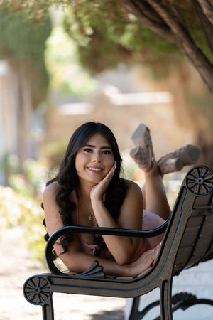 Young woman with long dark hair smiles while lying on an outdoor metal bench, resting chin on hand.