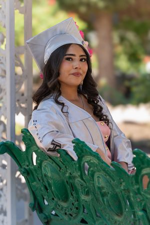 Young woman in white graduation cap and gown poses on green ornate bench outdoors.
