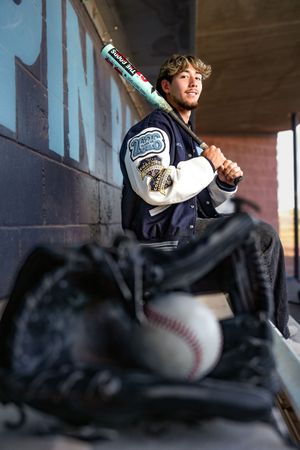Teen baseball player in varsity jacket holding bat on shoulder, with glove and ball in foreground of dugout.