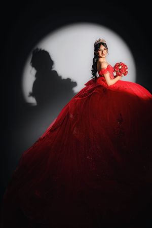 Young woman in elegant red ball gown with crown holds bouquet, posing dramatically in spotlight against dark background.