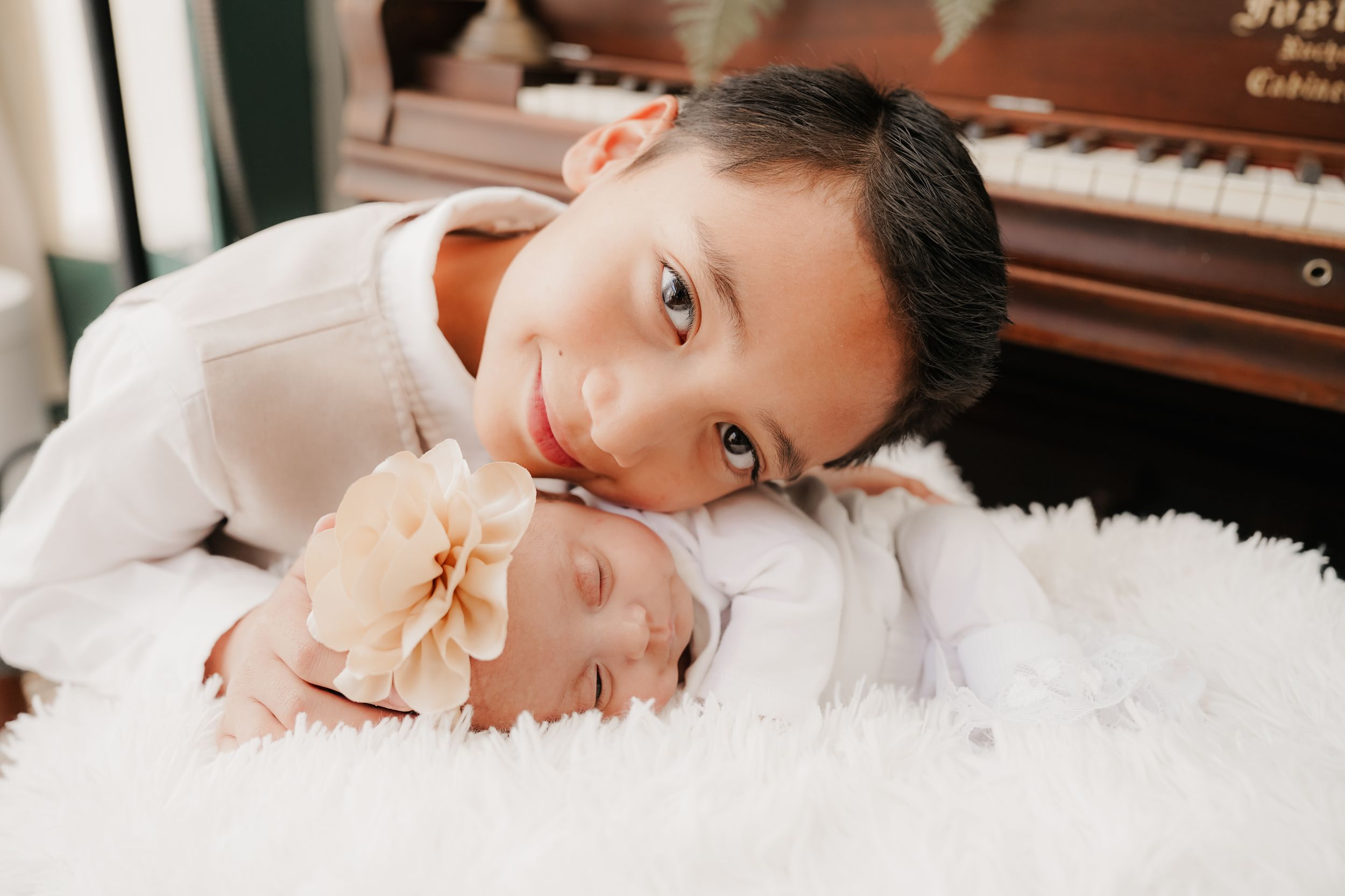 A smiling person in white clothing rests on a fluffy white rug next to an upright piano.