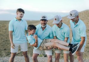 Group of males in matching mint green polos and white shorts posing outdoors, with one being carried playfully.