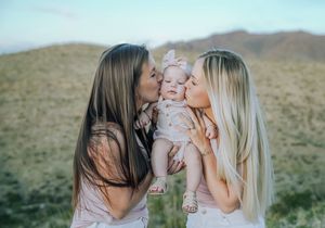 Two women with long hair kissing a smiling baby girl outdoors with green hills in the background.