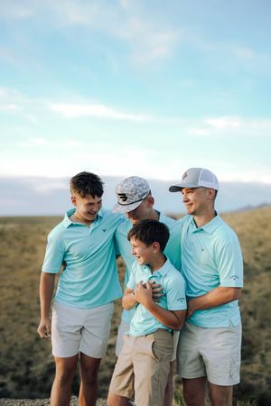 Four young men in matching teal polo shirts and light shorts pose together outdoors on a sunny day.