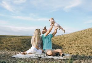 A young family sits on a blanket outdoors, with a man lifting a baby in the air while a woman watches joyfully.