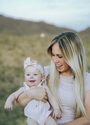 Smiling blonde woman holding a happy baby girl wearing a pink bow headband outdoors in a scenic field.