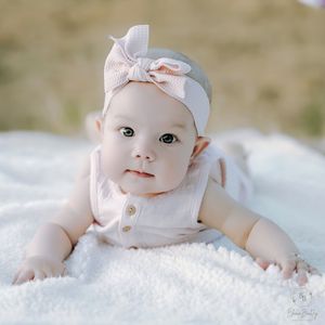 Adorable baby girl in white outfit and bow headband doing tummy time on a soft white blanket outdoors.