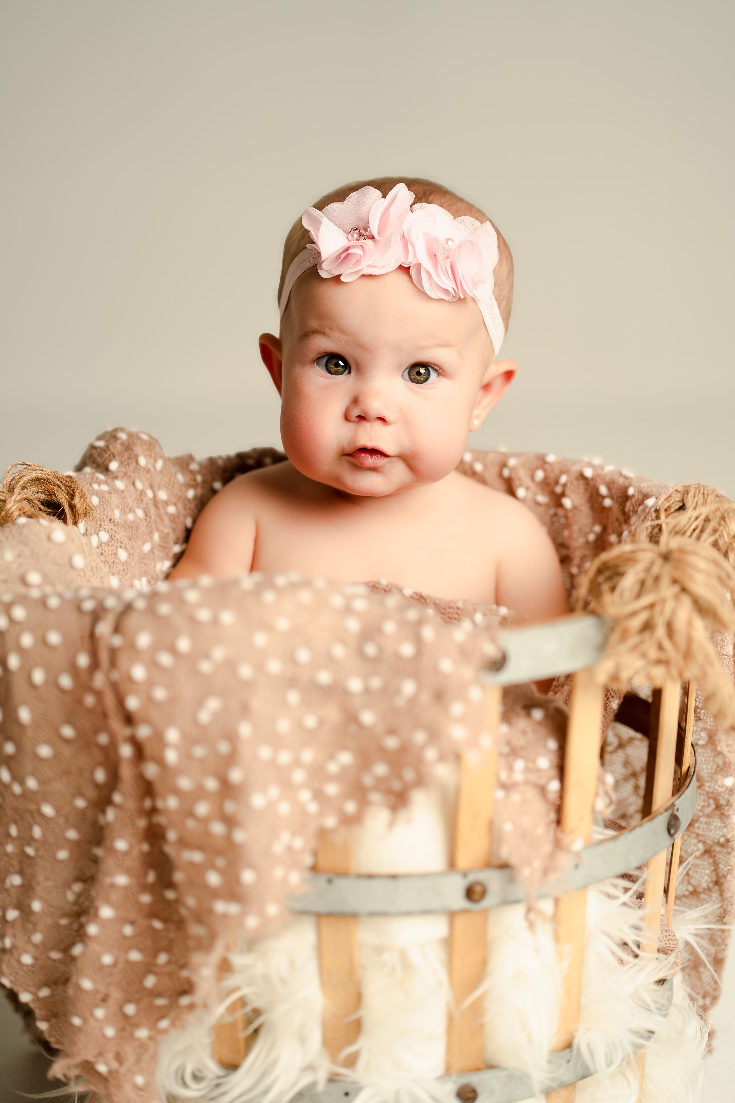 Adorable baby girl wearing a pink floral headband sitting in a rustic wooden basket with soft beige polka dot blanket.