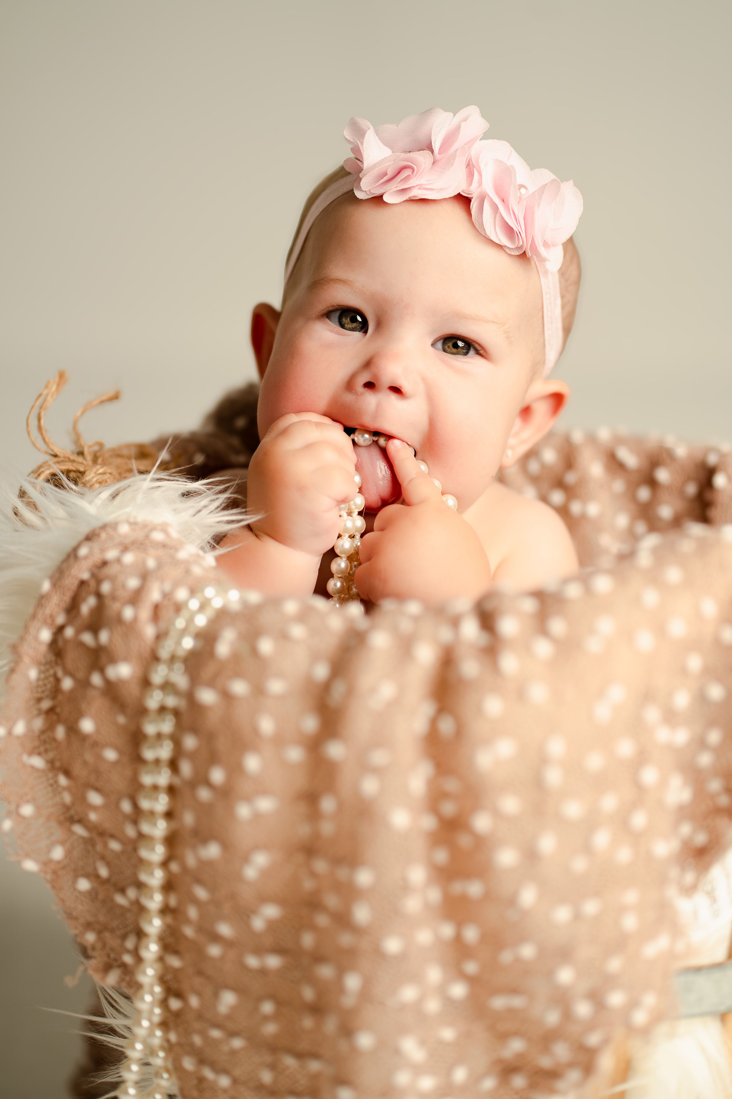Adorable baby girl wearing a pink flower headband sits in a basket chewing pearl beads, wrapped in a polka dot blanket.