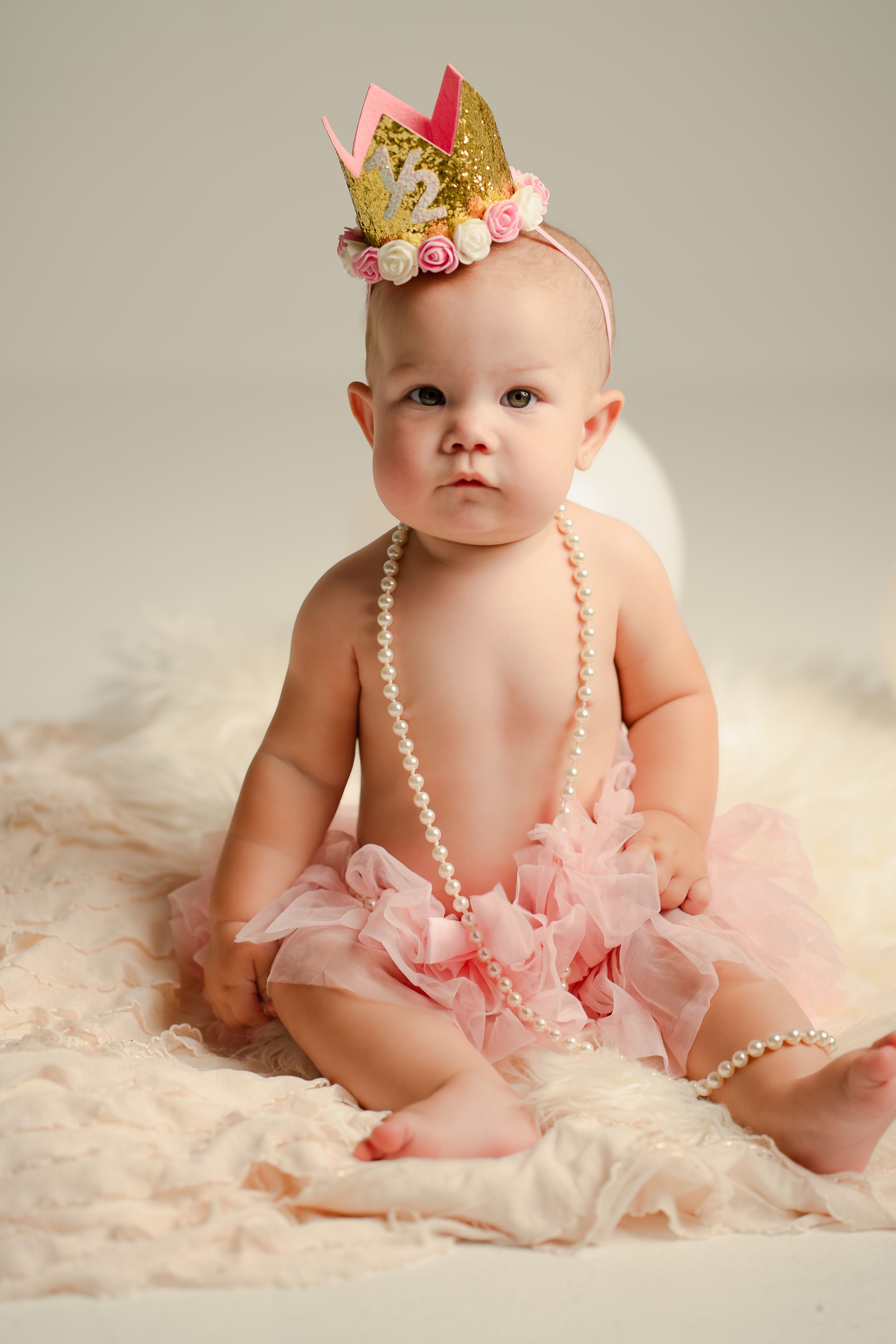 Adorable baby girl in pink tutu skirt, pearl necklace, and gold crown headband sitting on soft white blanket.