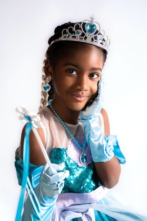 Young Black girl in teal sequin princess dress, tiara, gloves, and magic wand poses gracefully on white background.