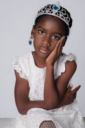 Young Black girl in white lace dress wearing blue crystal tiara and earrings, resting cheek on hand.