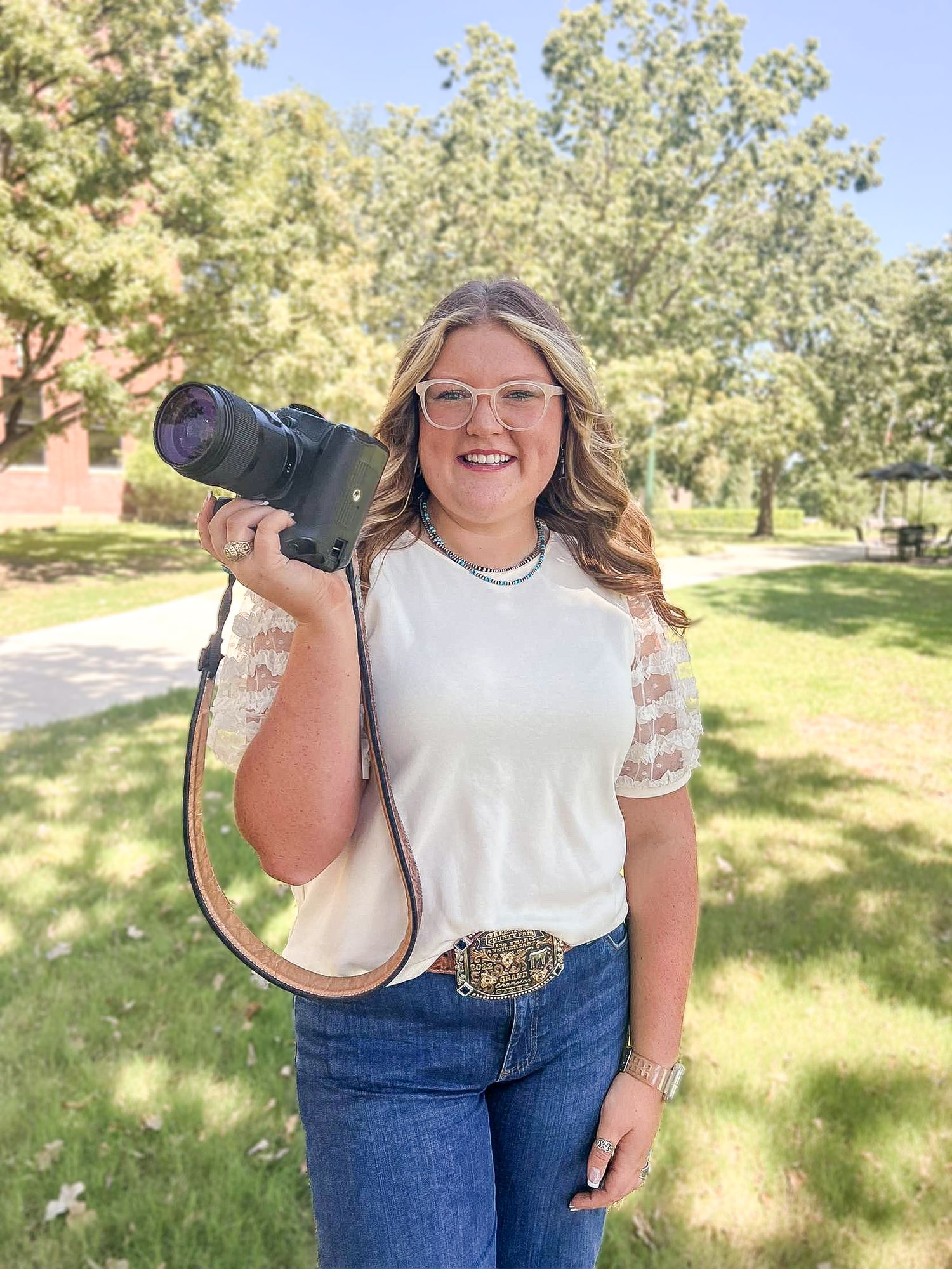 Smiling young woman photographer holding DSLR camera with telephoto lens outdoors on sunny college campus.
