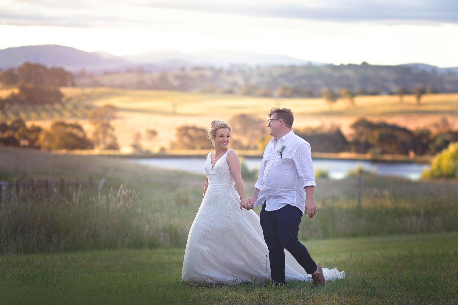 Bride and groom walking hand in hand through green fields with stunning golden sunset valley views.