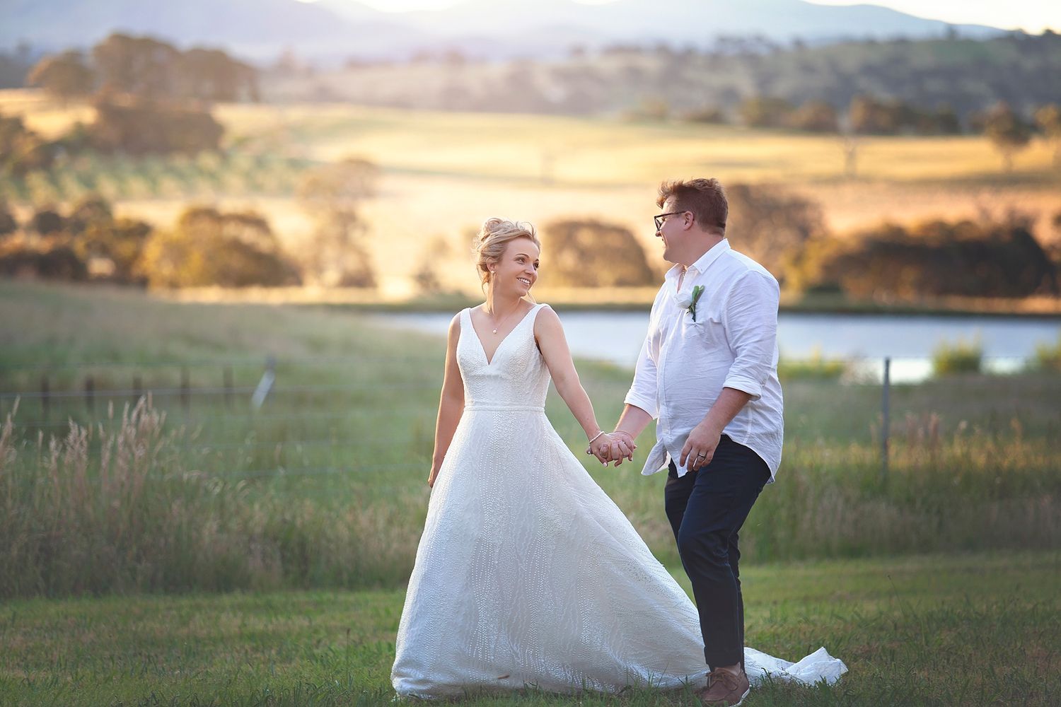 Bride in V-neck wedding gown and groom strolling hand in hand across scenic countryside at sunset.