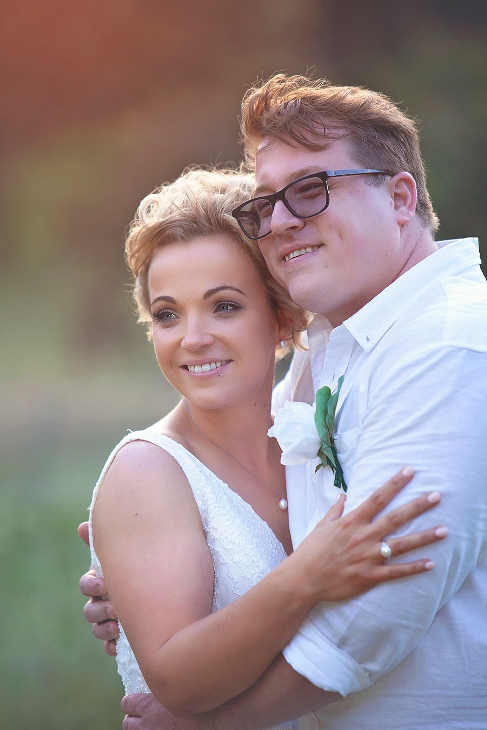 Smiling bride and groom portrait outdoors, groom wearing glasses and white shirt with boutonniere.