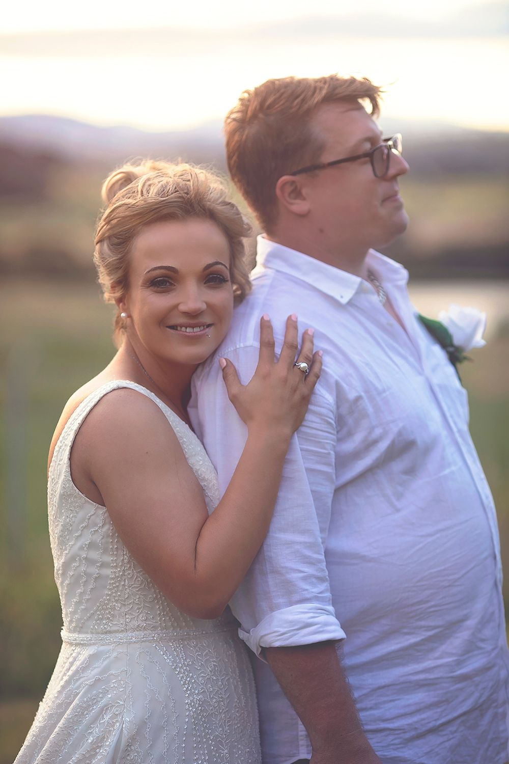 Bride leaning against groom's back smiling, both dressed in white with golden sunset countryside backdrop.