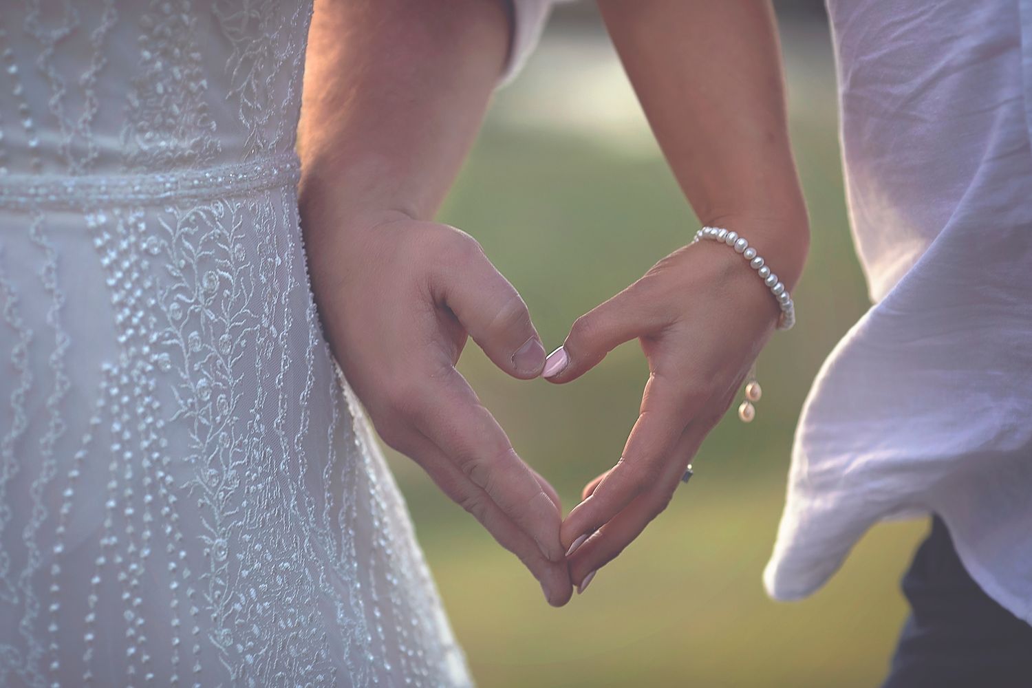 Bride and groom forming a heart shape with their hands, bride wearing pearl bracelet outdoors.