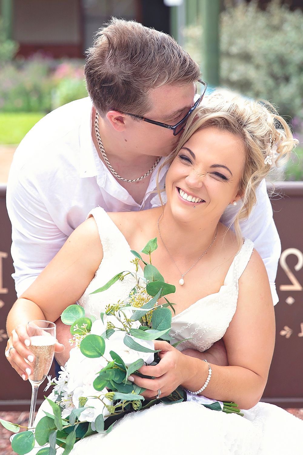Groom kissing bride's head as she laughs holding champagne and eucalyptus bouquet on a bench.