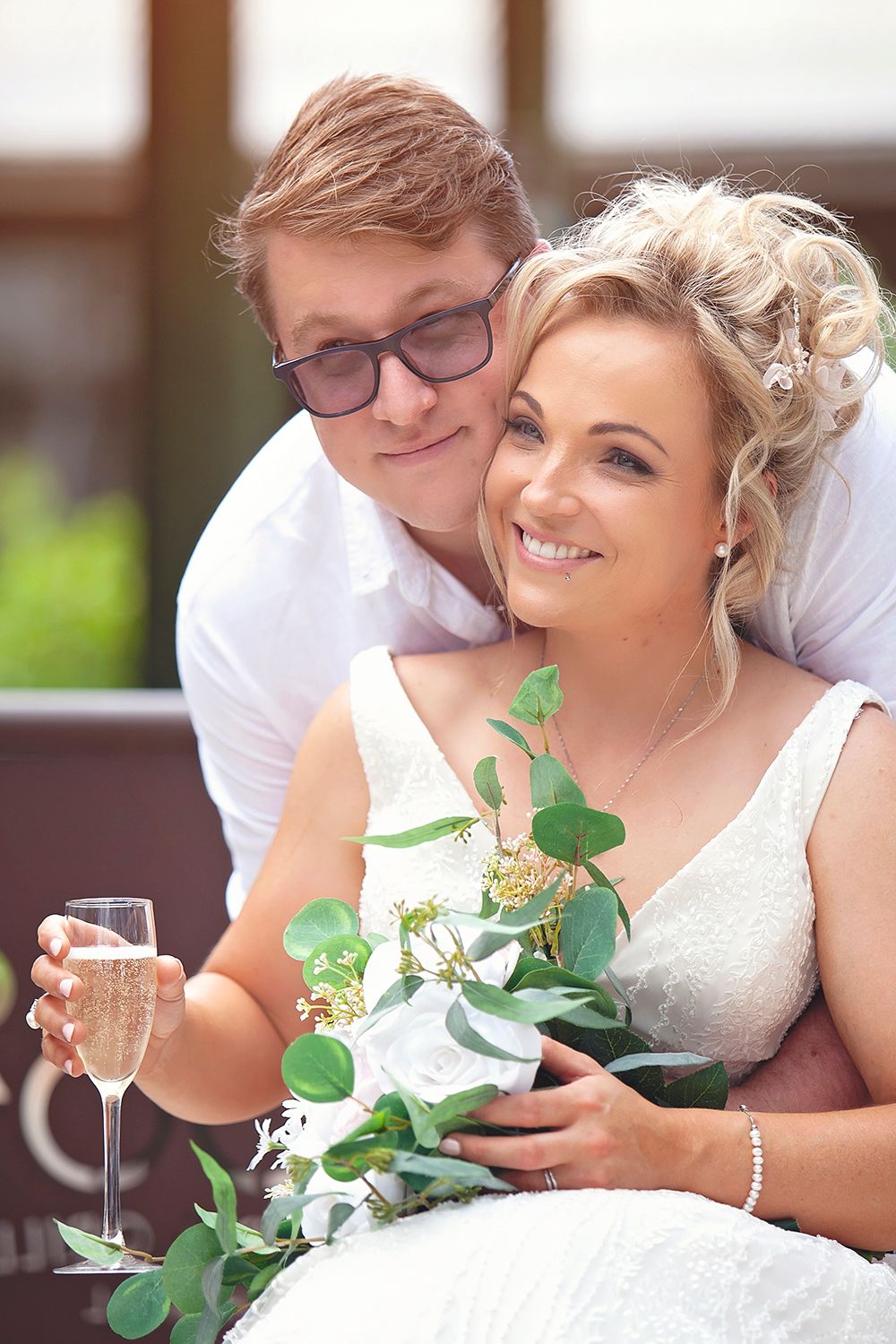 Smiling bride holding champagne and bouquet with groom embracing her from behind at wedding reception.