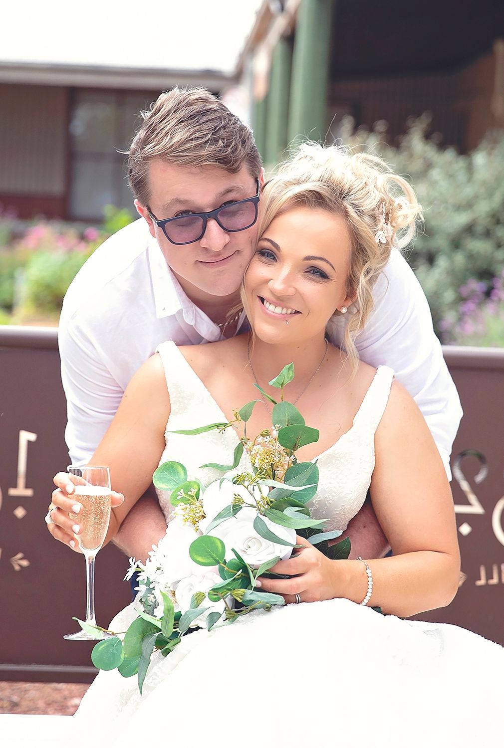 Happy newlyweds posing together outdoors, bride holding champagne glass and green eucalyptus bouquet.