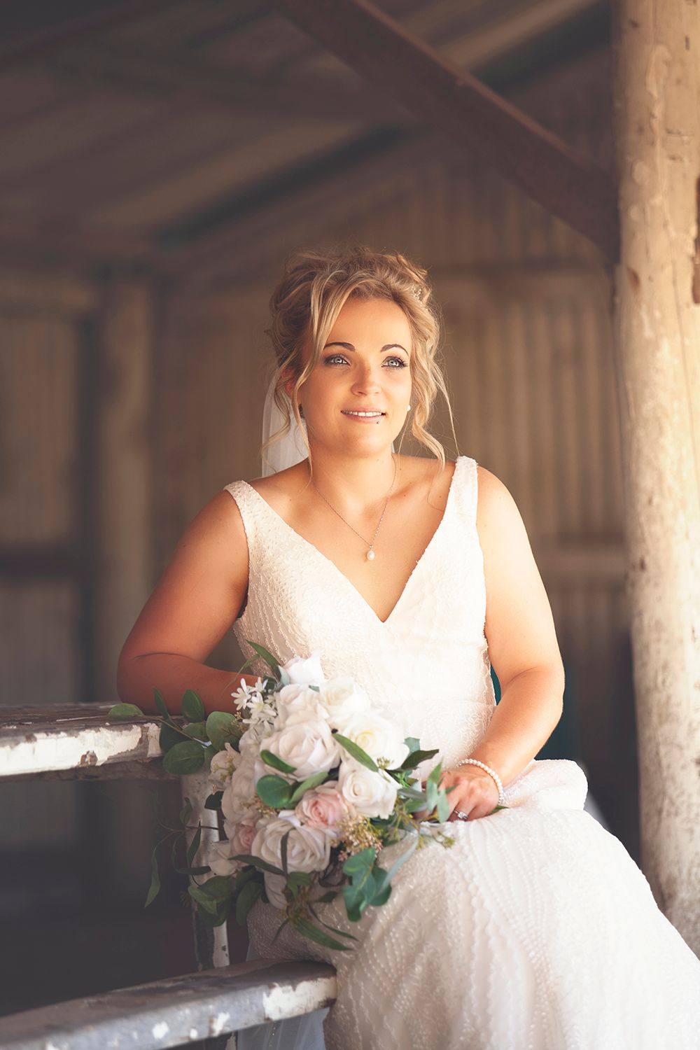 Elegant bride in white lace gown holding pink and white bouquet inside a rustic barn.