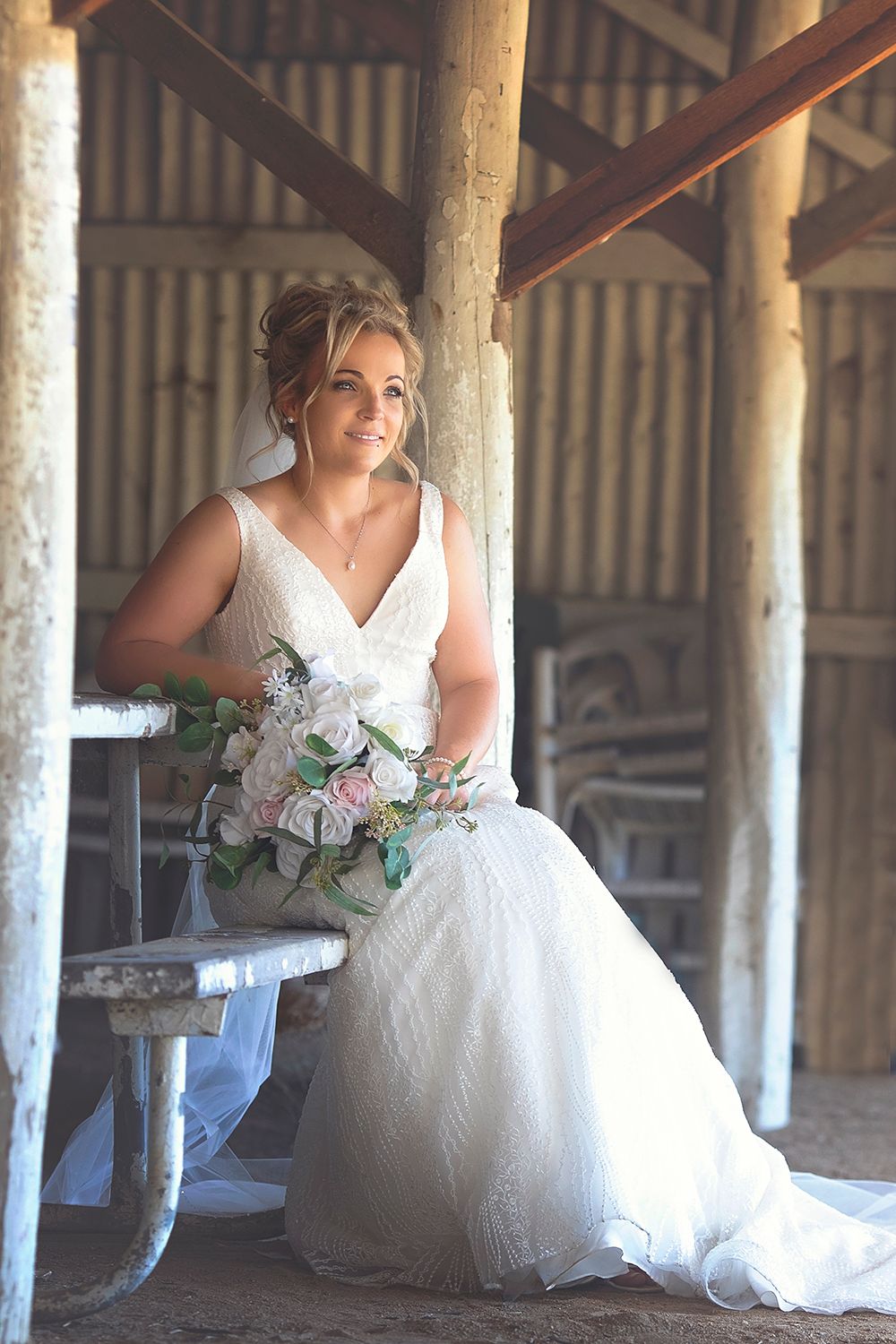 Beautiful bride in white wedding dress sitting in sunlit rustic barn holding floral bouquet.