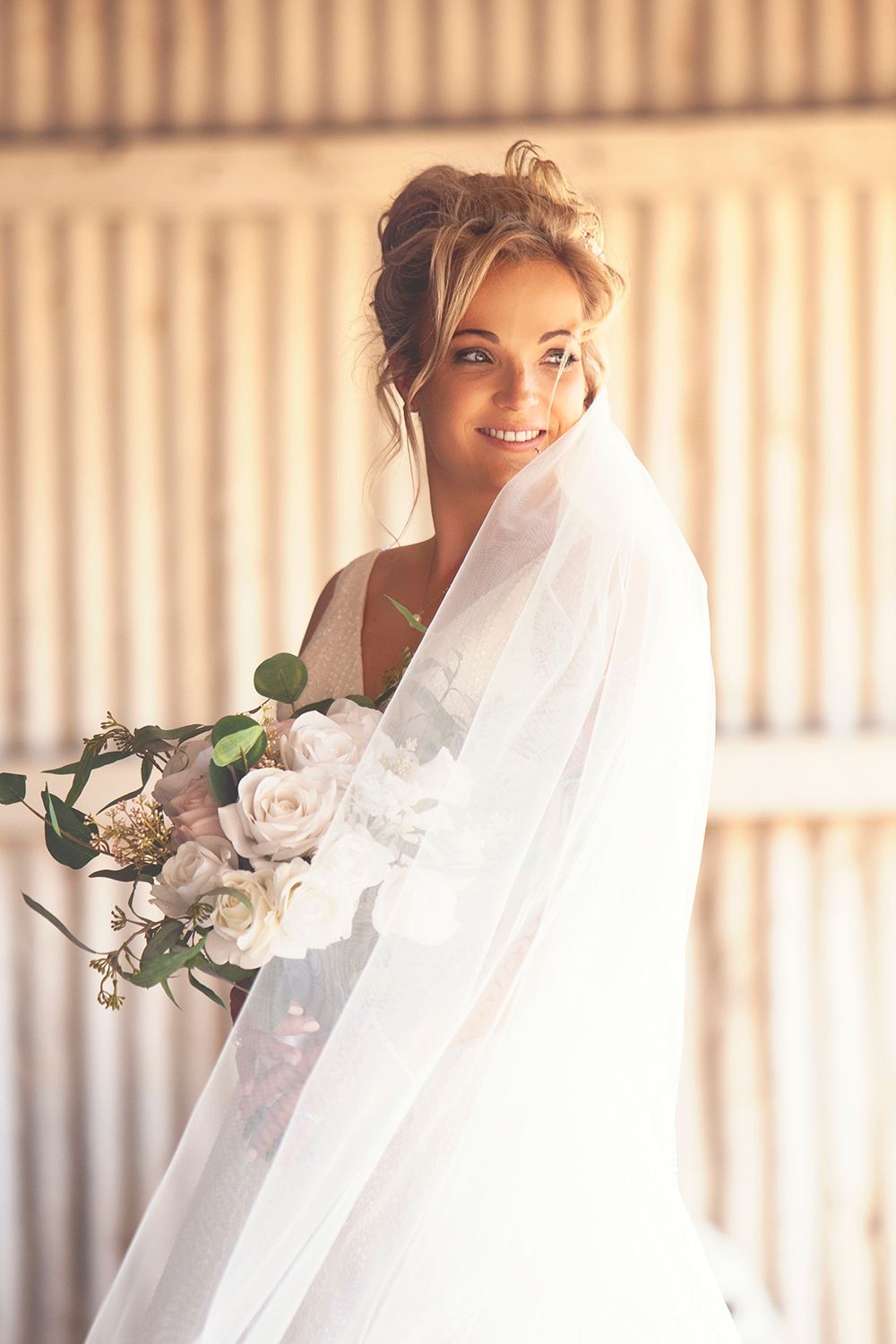 Radiant bride smiling with veil flowing, holding white rose bouquet inside a warm barn.