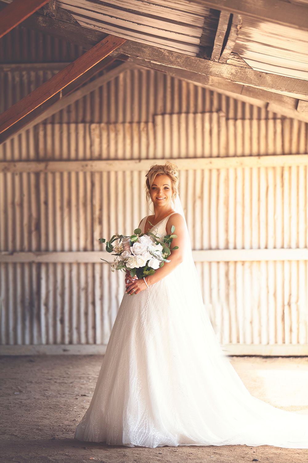 Bride in white tulle gown holding bouquet standing inside a sunlit rustic wooden barn.