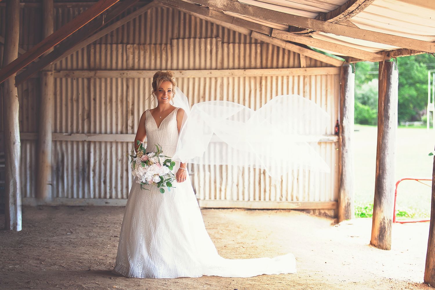 Bride in white wedding dress with veil billowing in wind inside a rustic barn.