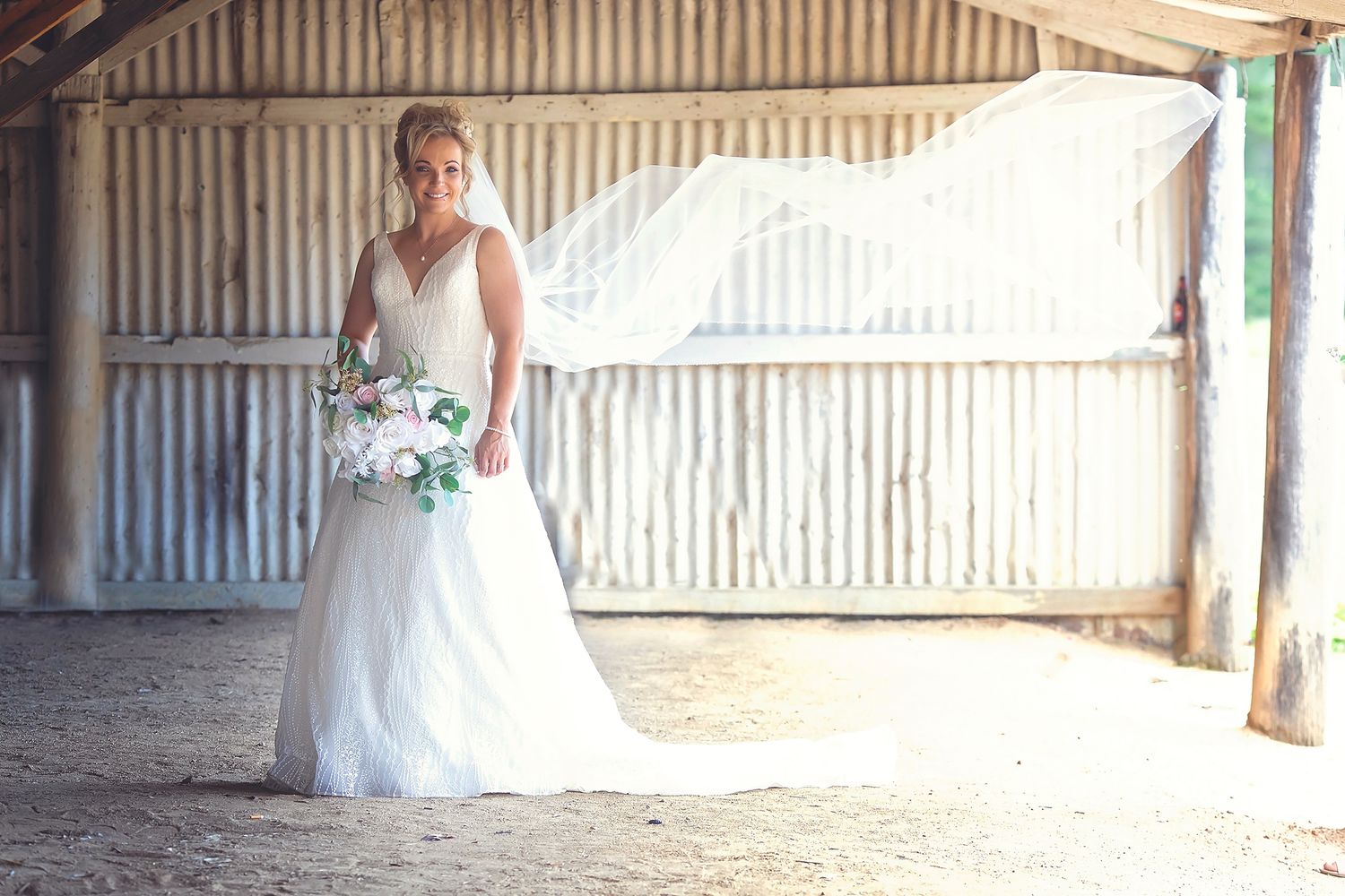 Bride in white wedding gown holding bouquet standing alone inside a rustic barn setting.