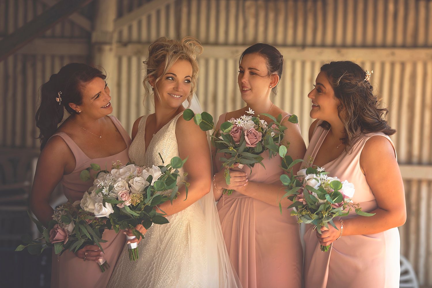 Bride with three bridesmaids in blush pink dresses holding white and green bouquets inside a rustic barn.