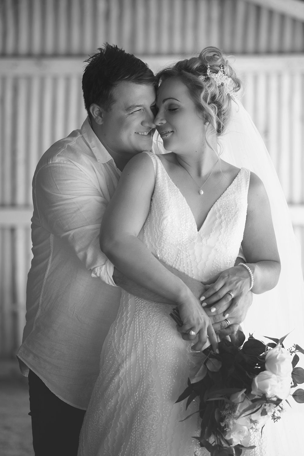 Black and white portrait of newlyweds embracing tenderly inside a rustic barn on wedding day.