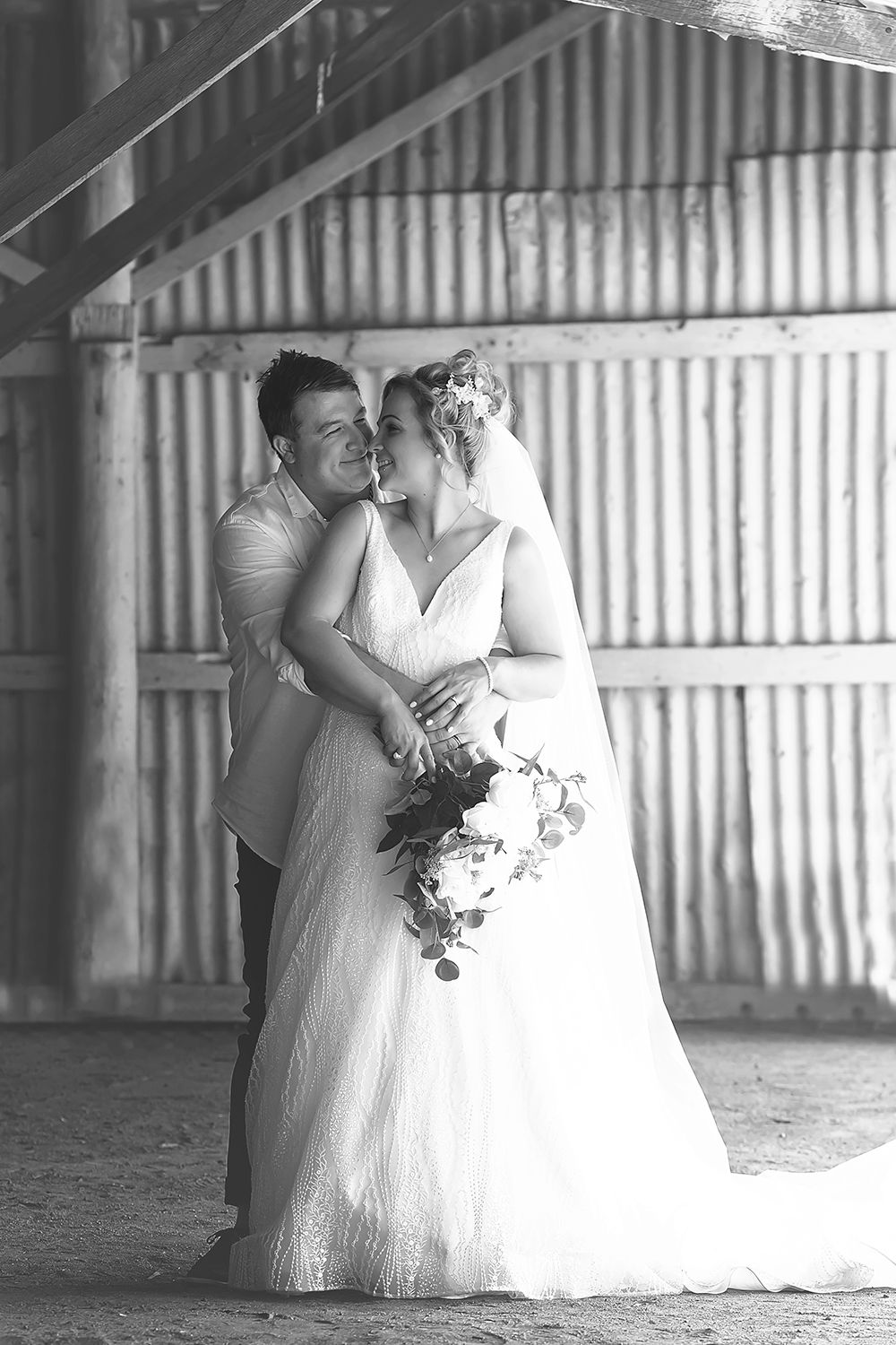 Black and white photo of groom kissing bride on cheek in a rustic barn wedding portrait.