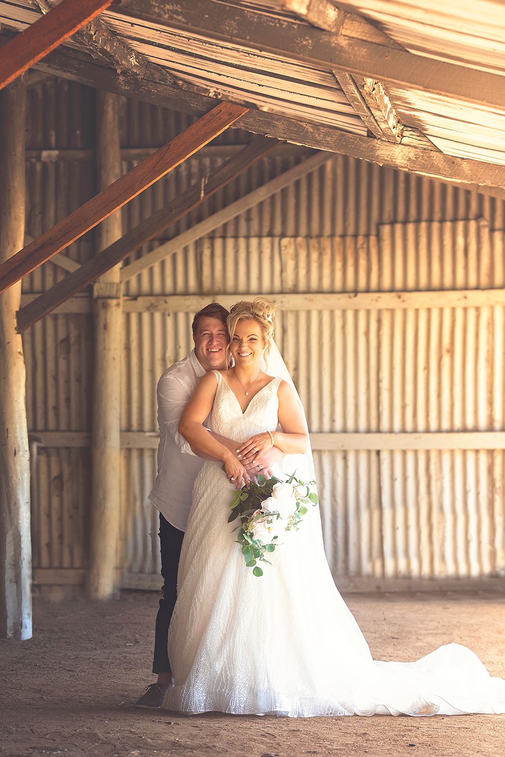 Bride and groom embracing inside a sunlit rustic barn, bride holding a white and green bouquet.