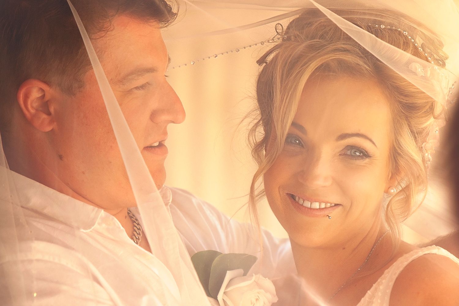 Close-up portrait of bride and groom sharing an intimate moment under a wedding veil in golden light.