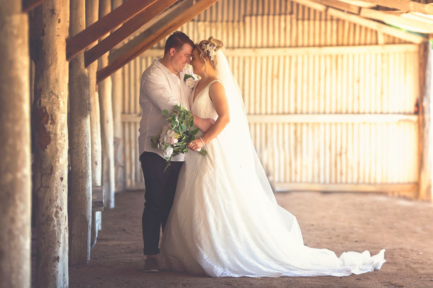 Bride and groom sharing a romantic kiss inside a rustic barn with wooden beams and corrugated walls.