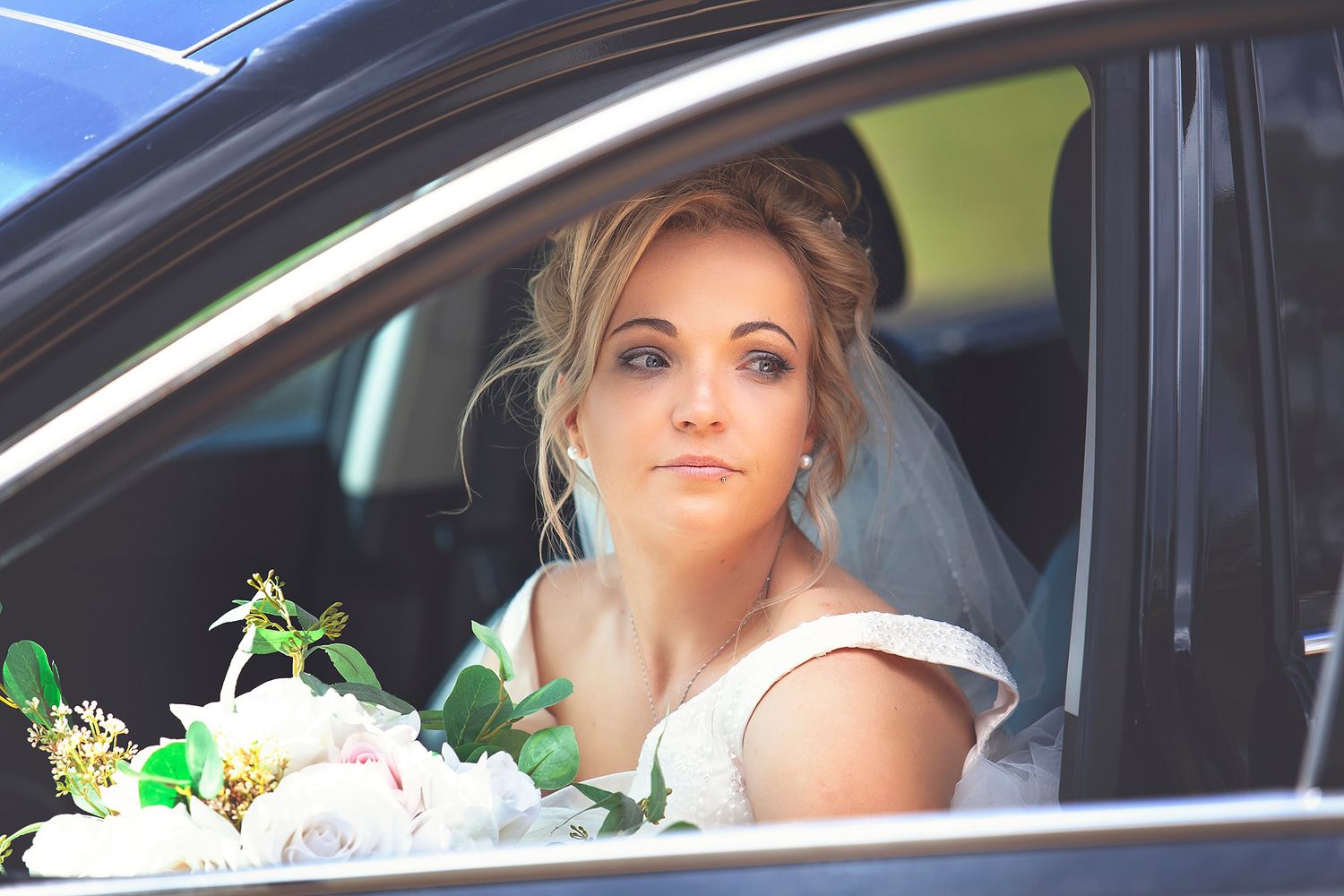 Elegant bride in white dress sitting in a car holding a bouquet of white and green flowers.
