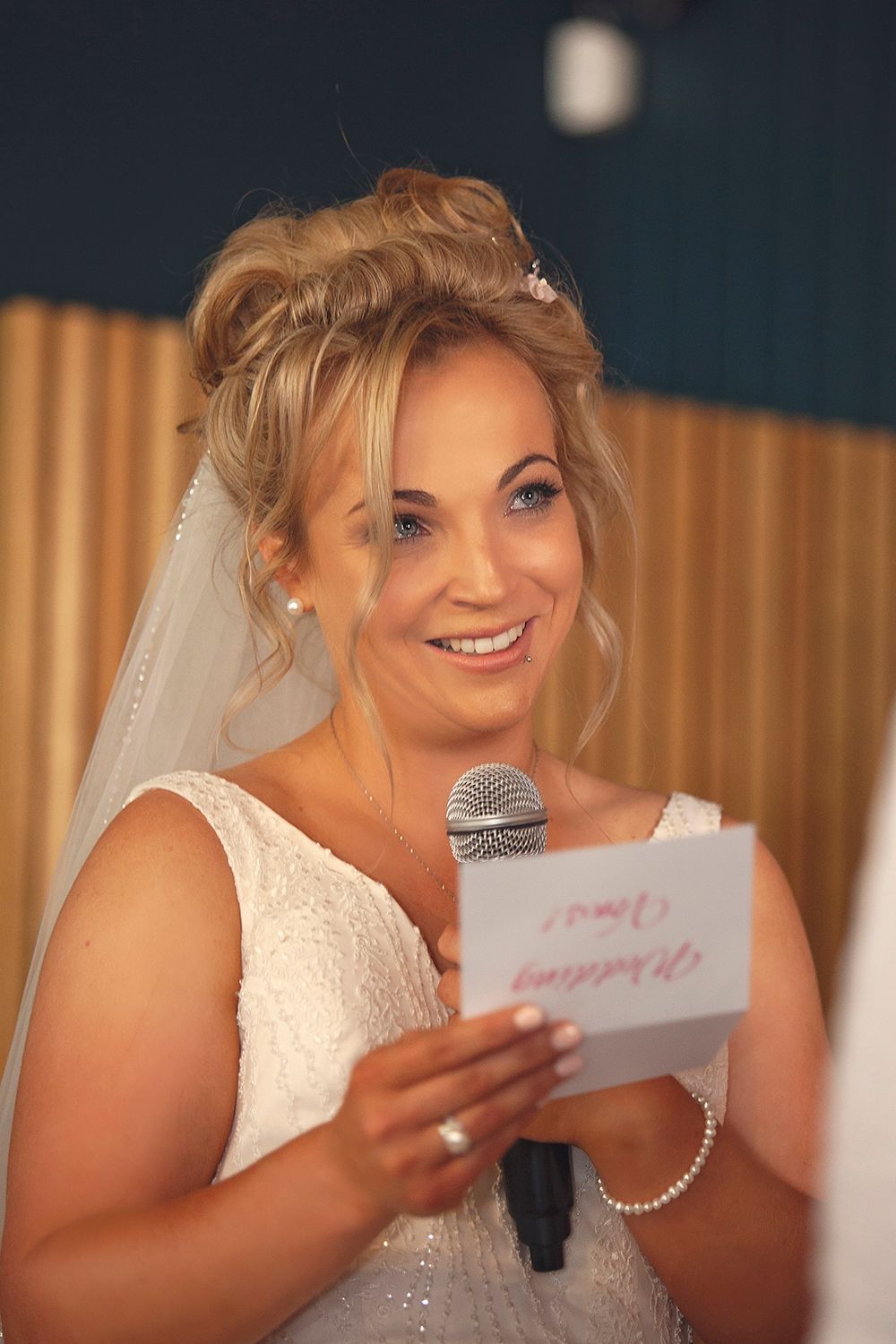 Smiling bride in white lace gown holding a microphone and reading wedding vows from a card.