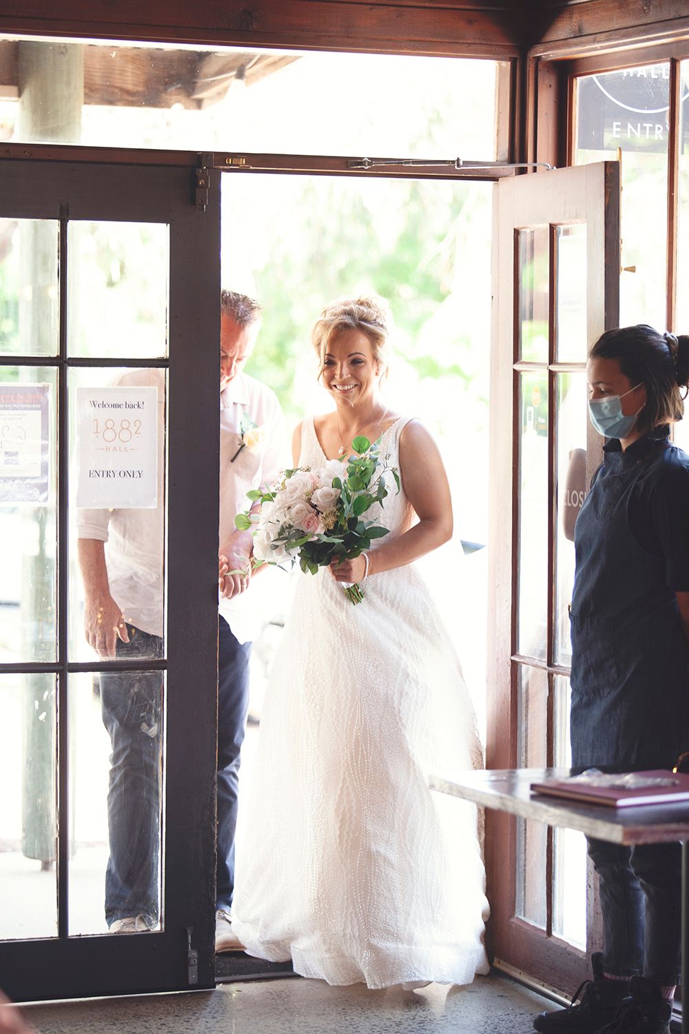Radiant bride in a white flowing gown entering a venue through glass doors holding a floral bouquet.