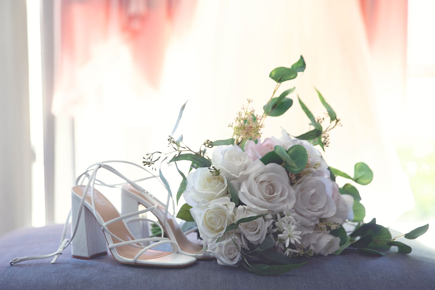 White bridal heels and bouquet of white roses with greenery on a table.