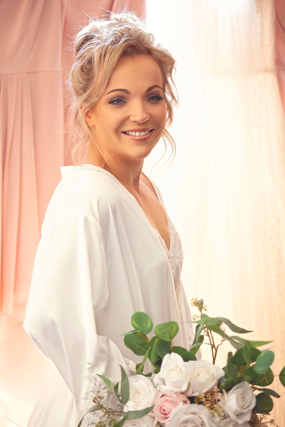 Radiant bride in white satin robe holding a bouquet of white and pink roses.