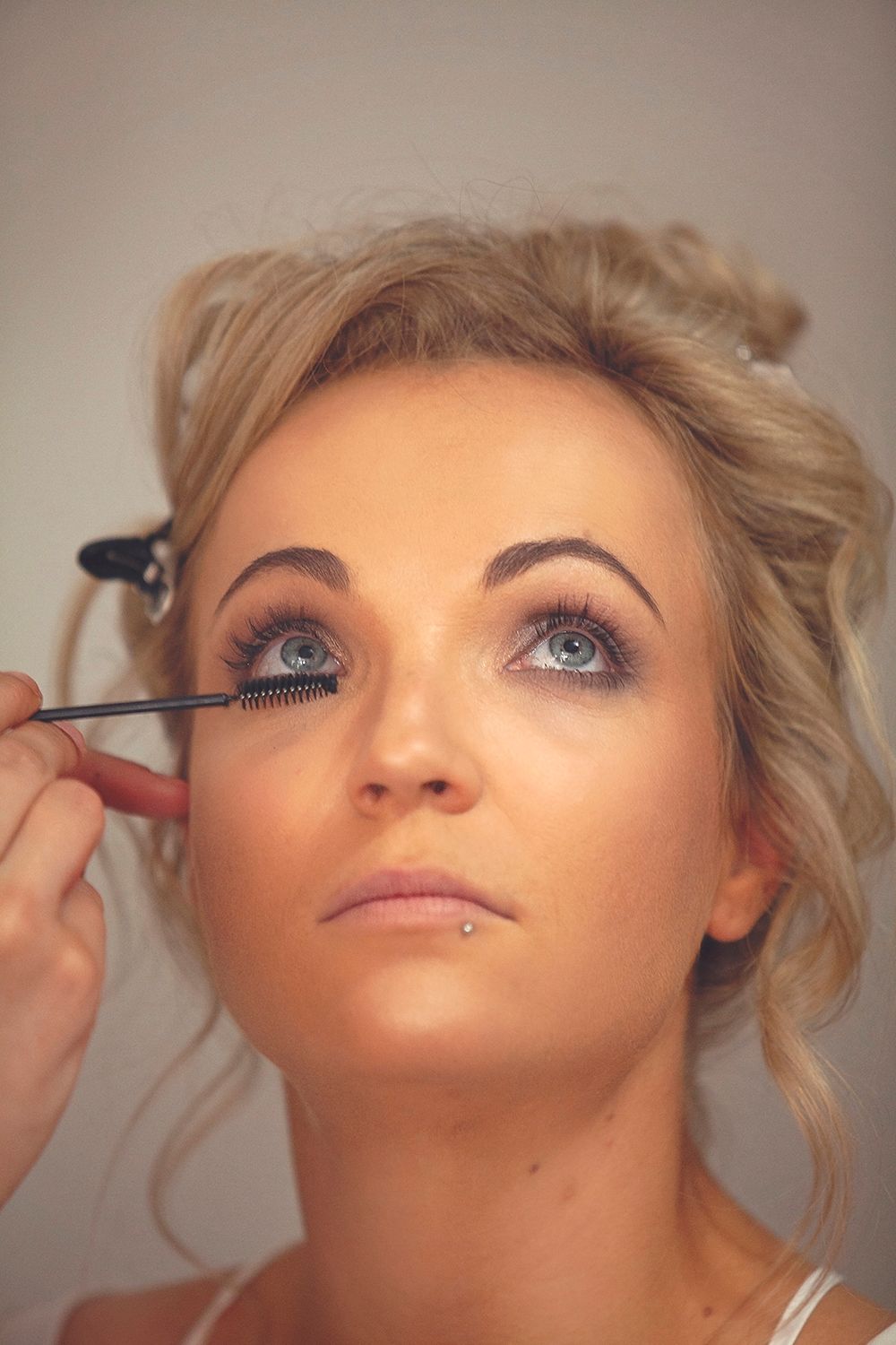 Blonde bride getting mascara applied by makeup artist during bridal preparation.