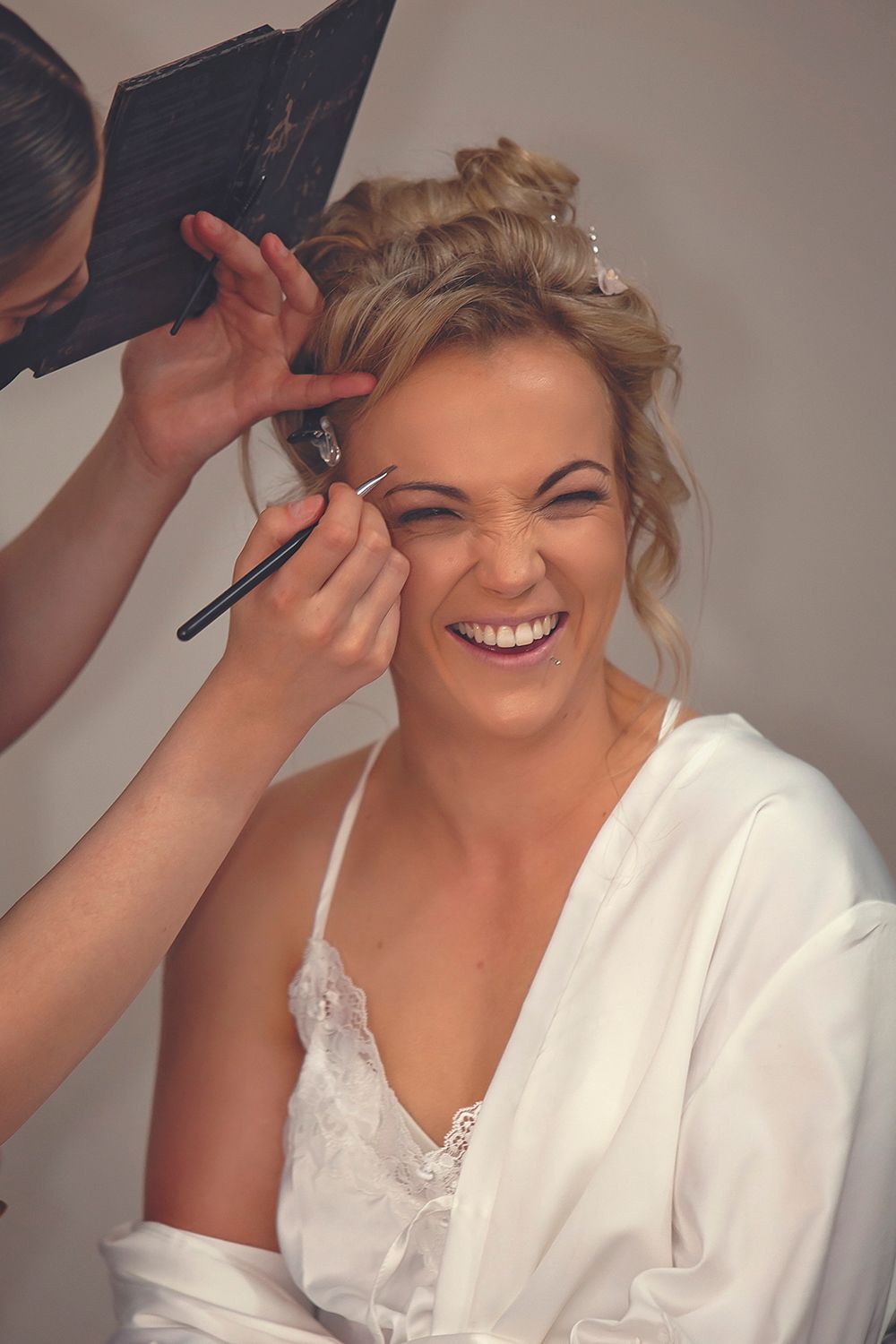 Laughing blonde bride in white robe getting eyebrow makeup applied before wedding.